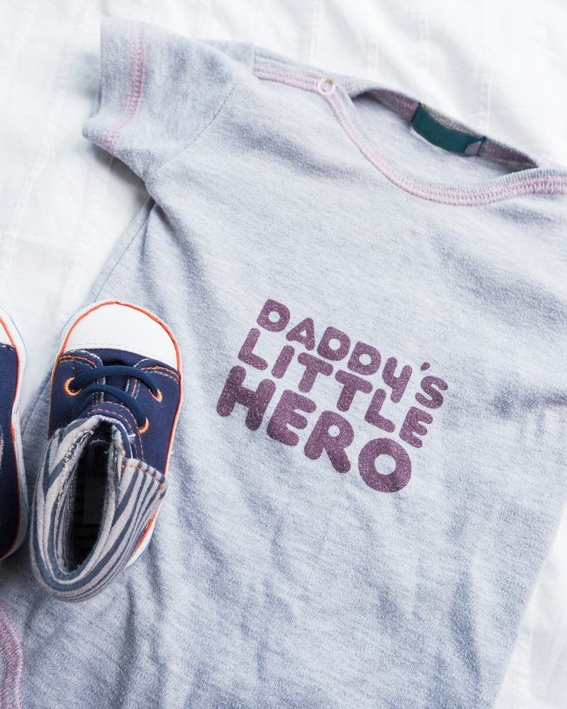 Grauer Babybody mit dem Aufdruck Daddy's little Hero und kleinen Schuhen